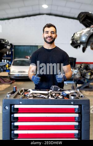 Cheerful male mechanic showing thumbs up while standing near tool cabinet with various instruments in car service and looking at camera Stock Photo