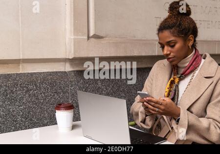 Stylish young African American female freelancer in trendy outfit talking on mobile phone and working remotely on laptop in cafe Stock Photo