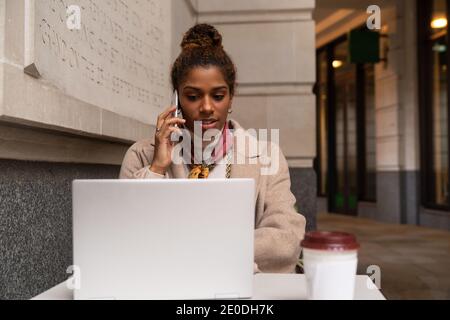Stylish young African American female freelancer in trendy outfit talking on mobile phone and working remotely on laptop in cafe Stock Photo