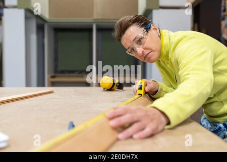 Low angle of concentrated middle aged female carpenter in uniform and protective goggles measuring wooden plank with roulette during work at table Stock Photo