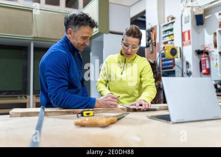 Concentrated adult male and female masters in uniforms measuring wooden board standing at table with laptop and instruments in modern workshop Stock Photo
