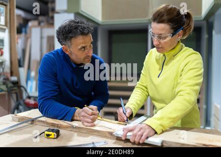 Concentrated adult male and female masters in uniforms measuring wooden board standing at table with laptop and instruments in modern workshop Stock Photo