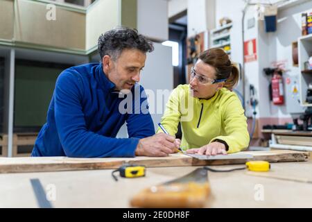 Concentrated adult male and female masters in uniforms measuring wooden board standing at table with laptop and instruments in modern workshop Stock Photo