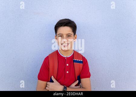 Adorable happy boy in red polo shirt standing near blue wall with backpack after school Stock Photo