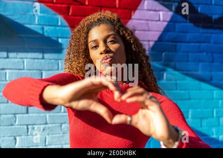 Confident young ethnic lady with Afro hair in casual wear pouting lips and showing heart sign with hands while standing on street near brick wall with Stock Photo