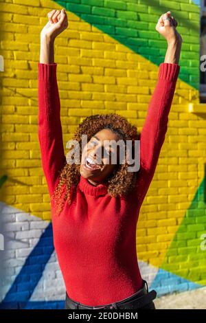 Excited young ethnic lady with long curly hair in casual clothes screaming with fists up while standing on street near colorful graffiti wall Stock Photo