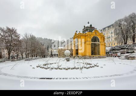 Marianske Lazne, Czech Republic - December 29 2020: The singing fountain in the foreground covered with snow and yellow Maxim Gorky colonnade house. Stock Photo