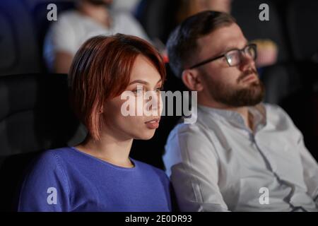 Selective focus of young couple watching movie in cinema, sitting in comfortable black seats. Side view of concentrated caucasian bearded man and woman with strong faces enjoying film. Stock Photo