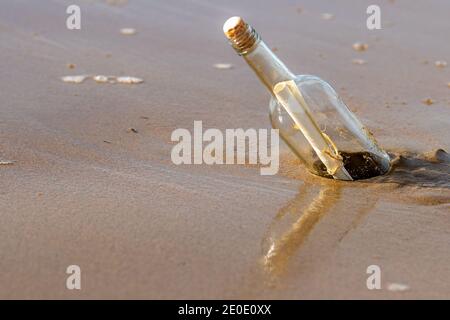 A message in a corked bottle washed up on a beach. The bottle is reflected in the wet sand below. The bottle is standing at an angle. Sunny day. Stock Photo