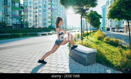 Fitness woman with perfect body stretching leg on bench, doing exercises after running workout in city streets. Stunning flexible woman warming up in rays of morning sun during jogging.