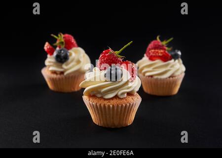 Three fresh tasty cupcakes with white creamy topping in row isolated on black background. Closeup front view of sweet delicious dessert with strawberry, blueberyy and rasberry slices. Stock Photo