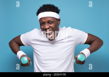 Strong young african fitness sports man doing exercise with dumbbells spend time in gym. Studio shot on blue wall. Stock Photo