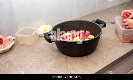 Close-up Of Preparing Traditional Jewish Cholent Hamin - Main Dish For The Shabbat. Raw Beef Being Added To A Saucepan Of Potatoes, Onion, Beans And Wheat Grains. Banner. Stock Photo