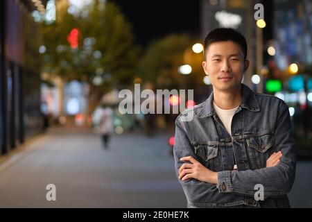 mid shot of one Asian young man at modern city at night, crossed arm and smiling, looking at camera Stock Photo