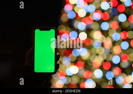 close up hand showing green screen phone with defocused colorful Christmas tree at dark night Stock Photo