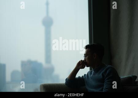 mid shot of one Asian young man sitting near window thoughtfully, blur modern landmark out of the window Stock Photo