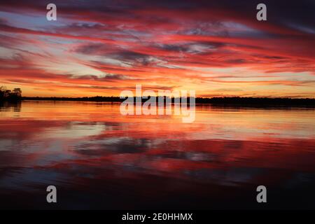 Sunset with mirror reflections on the surface of the water. Vibrant colors of red and golden swirls from the blue sky to tranquil tree-lined horizon. Stock Photo