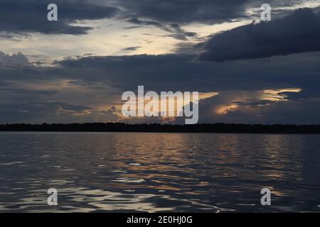 The calm before the storm as the dark gray clouds gather on the horizon. The last light reflects across the calm waters to a distant treelined shore. Stock Photo