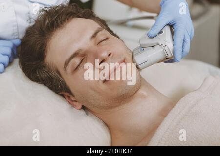 Attractive relaxed mature man getting non-surgical face lift. Professional cosmetologist using ultrasonic lifting machine on a male client. Handsome m Stock Photo
