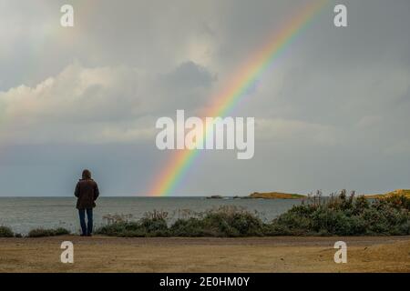 Man in hooded coat looking out across the Mediterranean sea towards a colourful rainbow Stock Photo