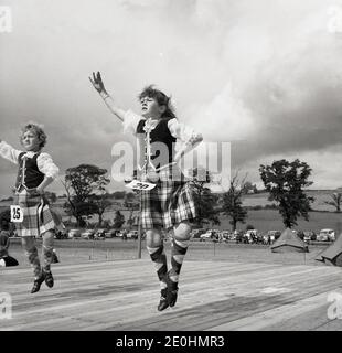 1960s, historical, two young girls dancing on a wooden platforn or stage outside in a competition at a Highand Games, Scotland, UK. Although the 'Aboyne dress', waistcoats, blouse and tartan skirt is worn for Scottish national dances, for competitive female Highland dancing, a kilt-based outfit as seen here is worn. Stock Photo