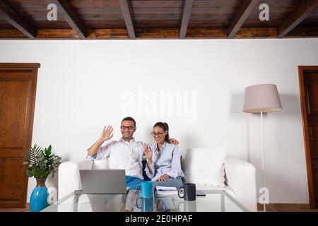 Young adult couple smiling, making a video call with their laptop in the living room at home. Stock Photo