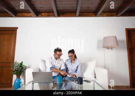 Young adult couple, taking notes, with the laptop, doing an online course, in the living room at home. Stock Photo