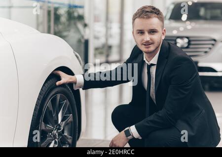 Professional car dealer examining car wheels and tires, working at the dealership salon. Young businessman choosing new car to buy at the automotive s Stock Photo