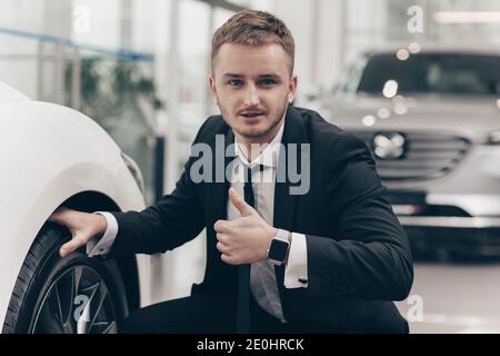 Professional car salesman smiling to the camera, showing thumbs up, posing near new automobile for sale. Car dealer examining wheel of an auto, showin Stock Photo