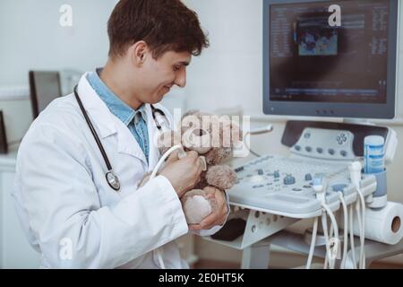 Handsome cheerful male doctor using ultrasound scanner on a plush toy teddy bear, copy space. Charming friendly pediatrician using ultrasound machine Stock Photo