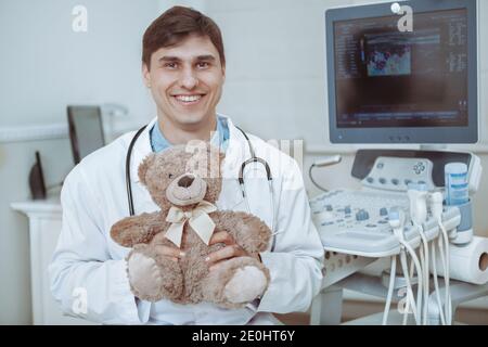 Handsome friendly male doctor holding plush teddy bear toy, smiling to the camera. Charming pediatrician holding teddy bear, waiting for medical appoi Stock Photo