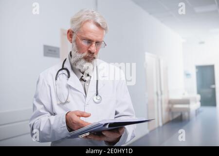 Professional medical worker reading documents on his clipboard, working at the hospital. Elderly doctor checking papers in the hallway of the clinic Stock Photo