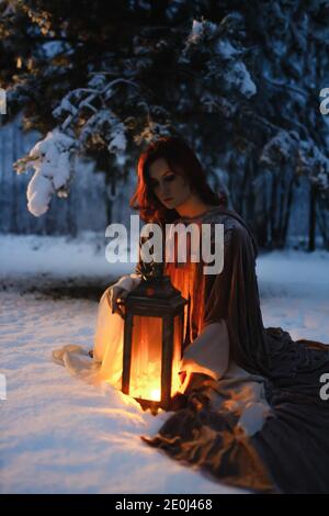 Young woman sitting under fir tree covered in snow looking at vintage lantern. Medieval fairytale concept Stock Photo