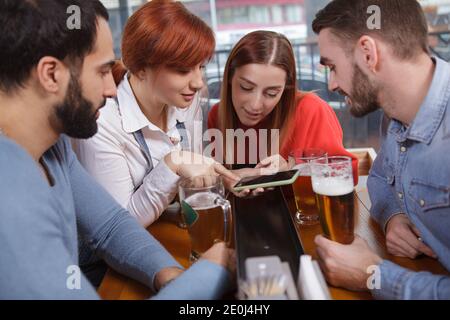 Group of friends using smart phone while drinking beer together. Young beautiful woman showing something online to her friends, using her phone Stock Photo