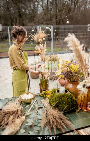 Young woman making compositions of dried and fresh flowers and herbs at the workshop outdoors. Florist, gardener or decorator composing floral decoration Stock Photo