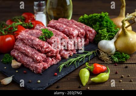 Raw minced meat cevapi ready for barbeque with various fresh vegetables on a wooden table. Selective focus. Stock Photo