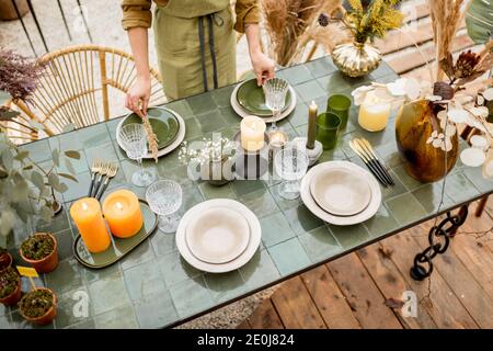 Young female putting herbs on a plates, decorating dinning table in natural Boho style in green tones outdoors Stock Photo