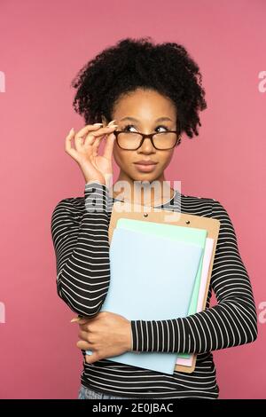 Afro-American tutor or teacher woman isolated on pink studio wall. Student girl touching glasses, holding notebooks, looking aside. Education Stock Photo