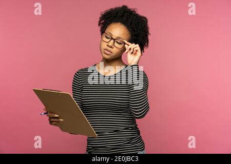 Thoughtful Afro-American student woman wear glasses holding folder, makes notes in document, thinking, isolated on pink studio wall. Planning concept. Stock Photo