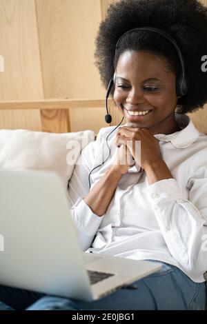 Smiling Afro-American millennial woman wearing headset, communicating via video chat on laptop, talking in zoom, watching webinar or video stream Stock Photo