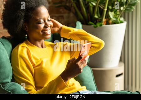 Close up of African American millennial young woman wear yellow sweater sitting in green chair, resting, using smartphone, typing message Stock Photo