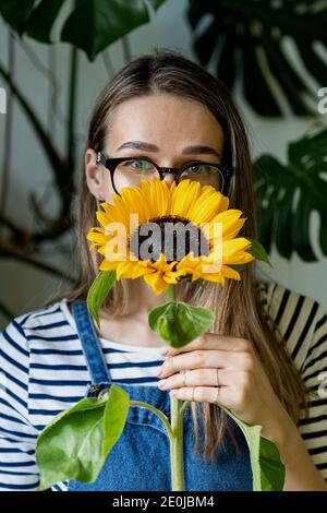 Florist woman in glasses holding and hiding behind a sunflower, looking at camera. Stock Photo