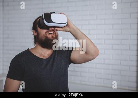 Bearded male hipster looking shocked with his mouth open, wearing virtual reality glasses, copy space. Young man using vr 3d glasses at work. Man tryi Stock Photo