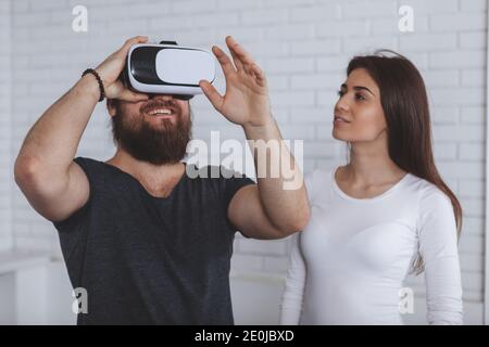 Bearded man using virtual reality headset, his girlfriend watching him. Young couple trying 3d vr glasses at work together. Friends using virtual real Stock Photo