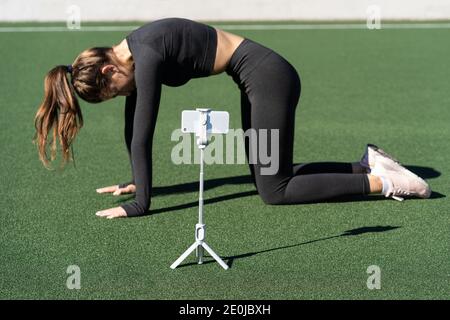 Woman doing yoga exercise called Cat pose outdoor, demonstrating exercises for her online blog, records on smartphone camera on tripod. Streaming a le Stock Photo