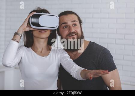 Cheerful bearded man and his girlfriend trying virtual reality glasses for the first time, copy space. Woman wearing 3d vr goggles, her biyfriend stan Stock Photo