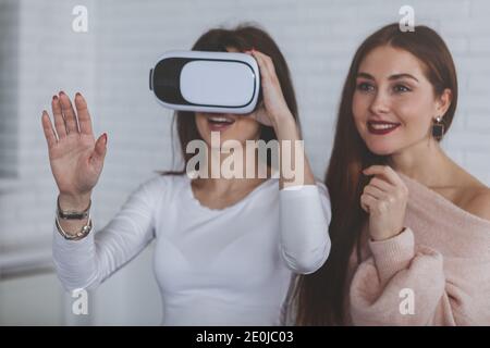 Two young women enjoying using virtual reality glasses together. Beautiful woman looking excited, her friend wearing 3d vr headset, copy space. Startu Stock Photo