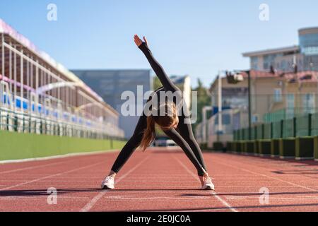 Young fit woman in black sportswear stretching legs on a treadmill rubber stadium on sunny summer day, standing forward fold posture, warming up befor Stock Photo