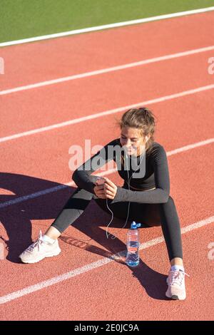 Fit woman in sportswear resting after workout or running, sitting on a treadmill rubber track, using and listens to music with wired headphones on sma