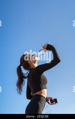 Thirsty young fit woman drinking water from plastic bottle, resting after jogging, holding mobile smartphone in her hand, listens to music. Blue sky w Stock Photo